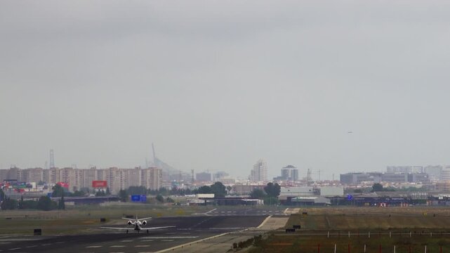Small private jet completing landing roll on runway, rear view, urban skyline with stadium structure and buildings visible through haze in background