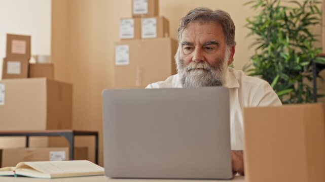 Man covers face with hands at cluttered desk with laptop, open notebook and stacked cardboard parcels and boxes inside a building; stress overwhelm.