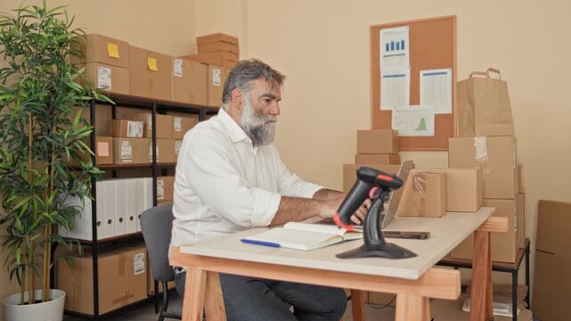 Man with beard stretches arms and leans back in chair at desk piled with parcels, laptop and barcode scanner in building; small business relief.