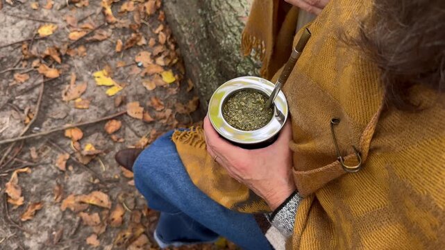 Overhead shot of a woman in a yellow poncho on a log, surrounded by fallen autumn leaves. She is holding a yerba mate cup with a metal straw called bombilla, a traditional South American infusion