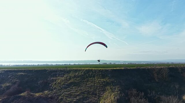 A person is paragliding above green hills next to a river. The weather is sunny with clear skies. The view shows a wide landscape and open space. The activity takes place during daylight.