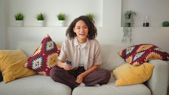 Young African American woman with curly hair sitting cross-legged on sofa, using a TV remote. Calm leisure lifestyle in a bright modern living room with colorful ethnic pillows and plants.