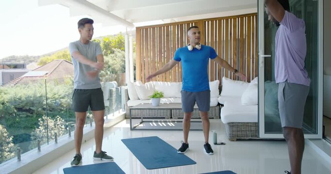 Diverse male friends following leader initiating arm swings, warming up on balcony with blue mats