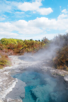 Rotorua, New Zealand: Vertical view of a vibrant blue geothermal hot spring pool with steam and white silica margins at Te Puia under a cloudy sky.