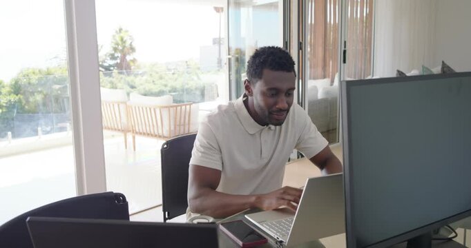 Mid adult African man starting work at desk, typing on laptop, checking monitor, referencing data