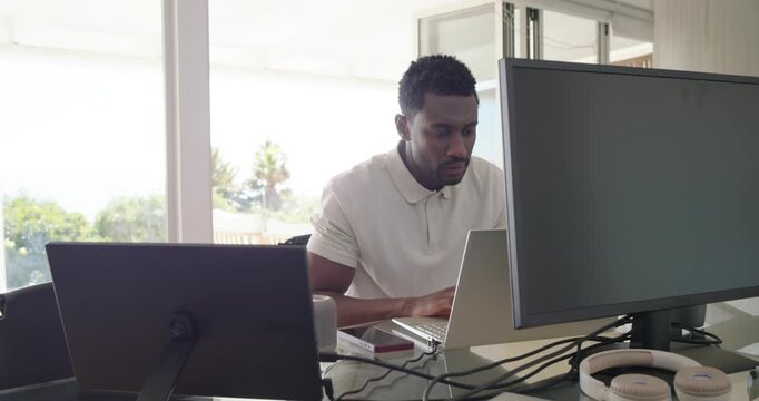 African American man in 30s at home office, seeing doc on laptop, typing and adjusting monitors