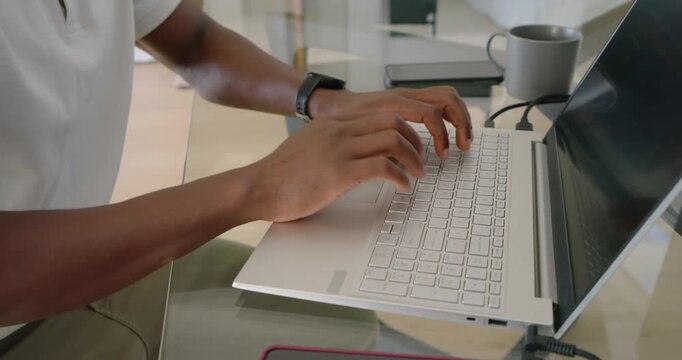 Mid-adult African man starting hands hovering, typing on white laptop at glass desk completing work