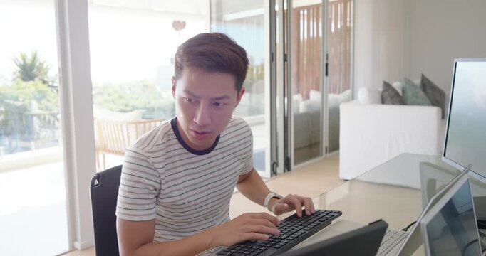Asian man typing on keyboard during remote work while using mouse and reacting to off-frame noise