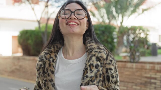 Woman wearing round glasses and leopard coat smiling with head tilted back and fingers near temples on a street; playful joy.
