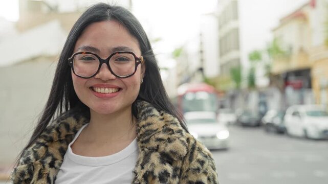 Woman with large round glasses and leopard coat smiling at camera on a busy city street with blurred cars and storefronts; confidence.
