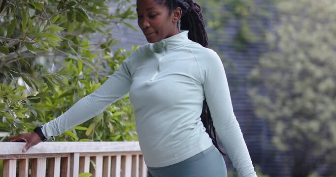 Bracing on deck railing adult African woman in sportswear holding right foot warming up, smiling
