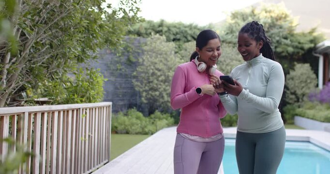 Diverse female friends reacting to ping and pointing at smartphone on poolside deck in activewear