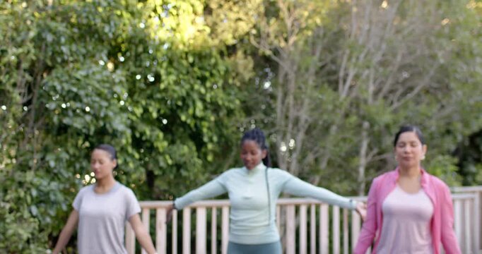 Diverse women doing yoga on deck after camera pull pressing palms overhead in activewear by railing
