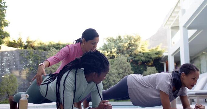 Hispanic instructor pink-top approaching fixing form two women holding forearm plank on patio mats