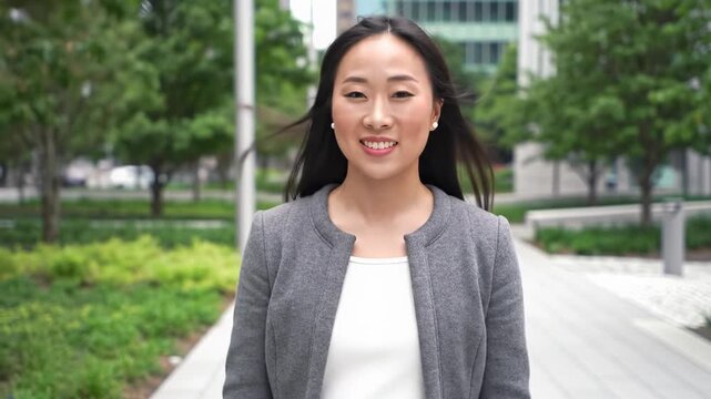 A confident young woman in stylish casual attire walks on an urban park path with blurred green foliage, a warm smile conveying approachability in bright daylight, slow tracking video, concept of