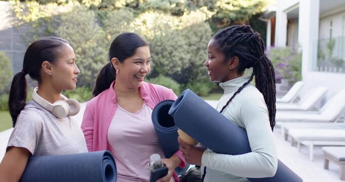 Diverse female friends greeting shifting blue mats and walking to yoga poolside in workout gear