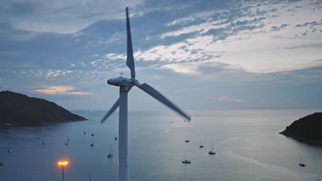 Wind turbine spinning its blades, generating renewable energy as yachts anchor in the bay of a tropical island at sunset, showcasing sustainable power and serene coastal landscape in Phuket, Thailand