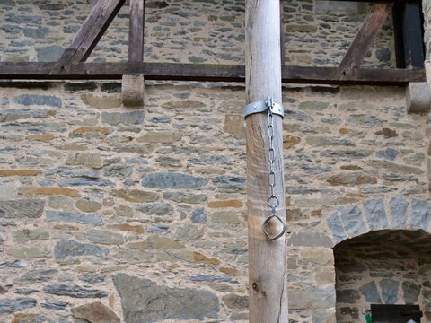 A close-up shot captures a historic iron pillory chain hanging from a vertical
weathered wooden pole. The metal shackle and loop, once used for public
punishment, are attached by a sturdy collar. In t