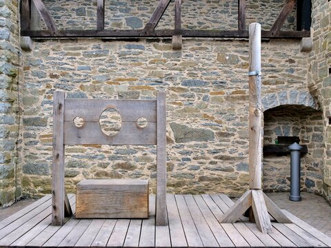 A wide shot displays authentic replicas of medieval punishment devices
arranged on a wooden platform. On the left stands a classic timber pillory with a
large head hole and two wrist cutouts, complete