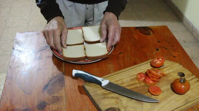 Persona colocando rodajas de pan lactal de miga sobre un plato para preparar un s&aacute;ndwich casero, junto a tomates reci&eacute;n cortados con chuchilla sobre una tabla de cocina. Escena culinaria cotidiana.
