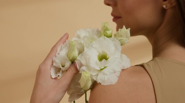 Close-up shot of lower face of young woman holding small bouquet of white elegant lisianthus flowers near her shoulder in studio