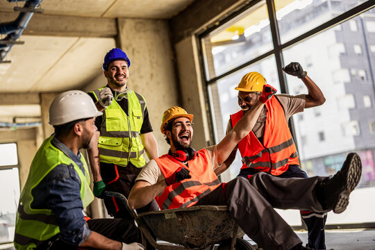 Cheerful construction workers playing with a wheelbarrow inside a building site.