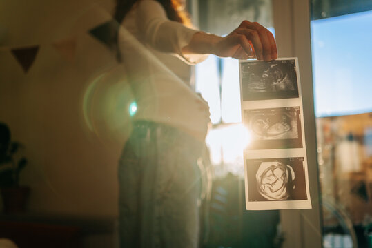 Pregnant woman standing in sunlit room, holding up multiple ultrasound scan prints, revealing developing baby images, feeling excited about impending parenthood and awaiting birth