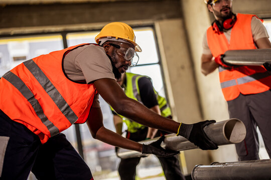 Builders in safety goggles and high-visibility vests lifting a large tube on site.