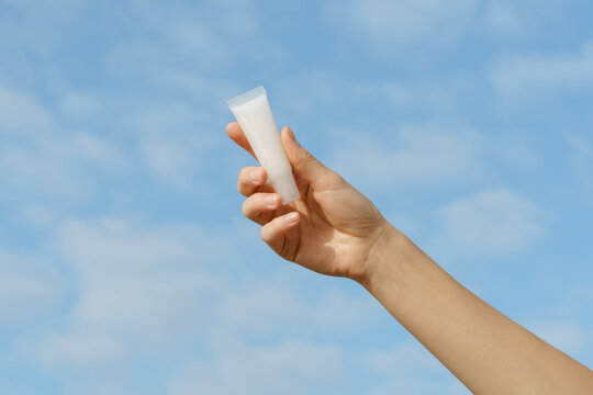 A female hand holds a blank white mockup cosmetic tube against a bright blue sky with clouds. Minimalist composition with natural light. Concept for skincare, beauty, sun protection, and natural produ