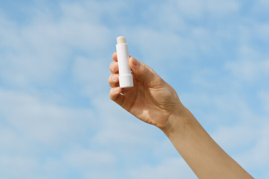 Woman's hand holds a blank white mockup lip balm against a soft blue sky. Minimalist scene with natural light. Concept for skincare, beauty, sun protection, cosmetic mockup, and self care outdoors