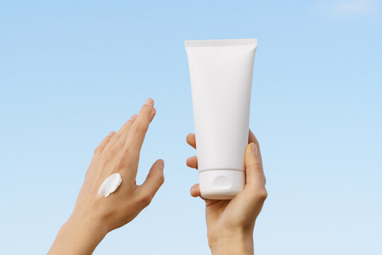 Hands hold a white cosmetic tube mockup, applying sunscreen or lotion to skin. The clear blue sky background suggests summer, skincare, and sun protection. Minimalist