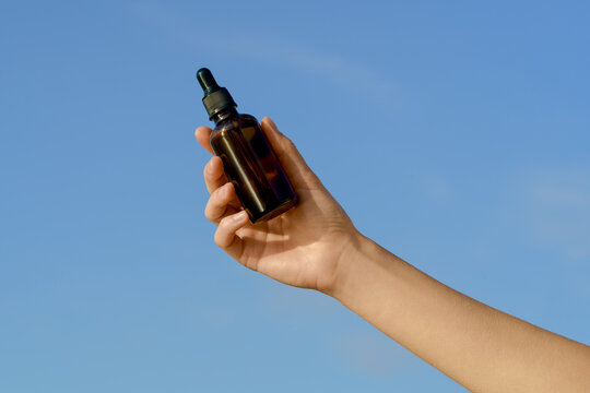A hand holds a brown glass dropper bottle up to a bright blue sky. Minimalist scene with natural light, ideal for cosmetic, beauty, or wellness concepts. Plenty of clean copy space