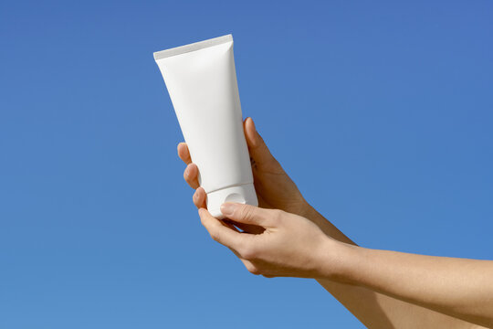 Female hands hold a blank white mockup cosmetic tube against a clear blue sky. Bright natural sunlight creates a clean, minimalist look. Ideal for skincare, lotion, or sunscreen product mockups