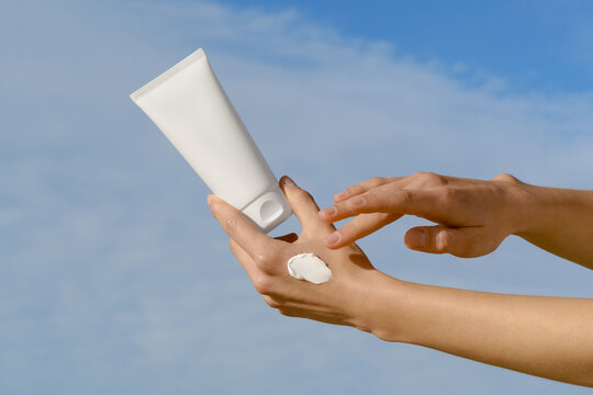 Woman's hands applying white cream from a blank cosmetic mockup tube against a blue sky background. Minimalist skincare concept with natural daylight. For sun protection, beauty, and wellness products