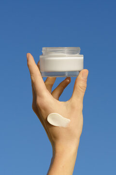 Hand holds a moisturizer mockup jar with a cream swatch on skin. Minimalist product shot with bright daylight against a blue sky. Concept for skincare, beauty, self care, and sun protection
