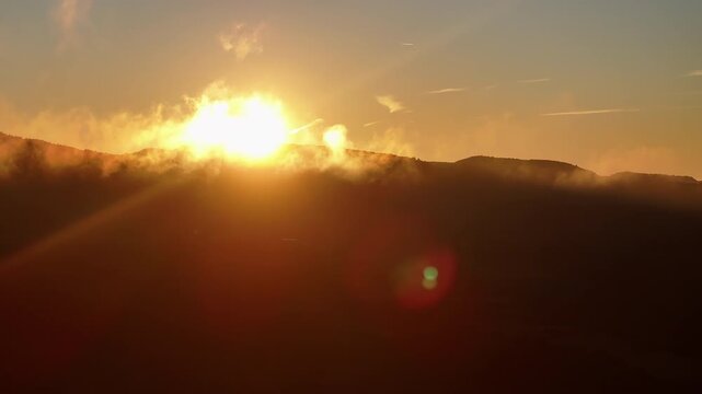 Sun glowing above a dark mountain ridge, warm orange tone saturating the sky, wisp of mist clinging to hillside and lens flare crossing the lower frame