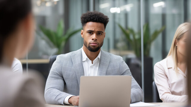 Business meeting with young male professional listening across a laptop, blurred colleagues in foreground and background, modern office consultation, ultra-realistic corporate phot