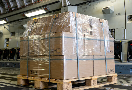 A stacked pallet of cardboard boxes wrapped in clear plastic inside a large aircraft cargo hold.