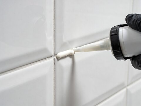 A gloved hand uses a caulk gun to apply white sealant into the joints of white glossy wall tiles.