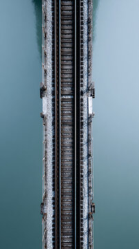Aerial view of a railway bridge spanning over calm water, showcasing parallel tracks and gravel bed, with reflections visible in the serene surface below