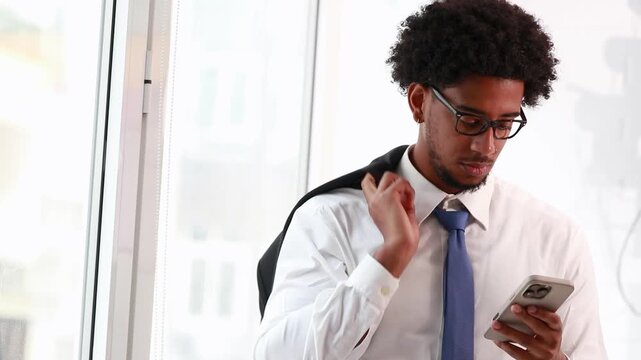 young man in a white shirt and blue tie puts on his suit jacket and looks at his phone by a bright office window
