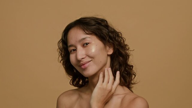 Portrait of positive brunette Caucasian gen Z girl smiling for camera after applying under eye patch against plain background in studio