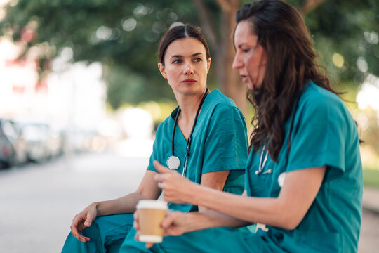 Two women in medical scrubs and stethoscopes enjoying a coffee break, sitting outside and having a serious conversation