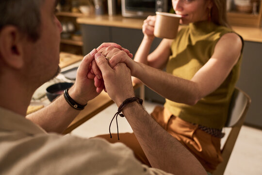 Couple holding hands across table while woman drinking coffee, showing intimacy and support during home conversation. Useful for relationship counseling or dating content