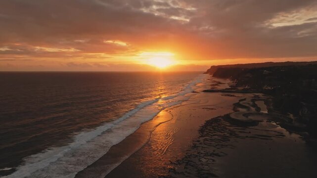 Golden sunset over the tranquil ocean at Melasti Beach, Bali, creating a vibrant orange reflection on the water and wet sand as white waves roll onto the tropical coastline. Aerial view drone flight