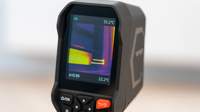 Handheld thermal camera displays temperature readings. Screen shows color-coded heat patterns. Yellow and red hues indicate varying temperatures. Digital display reads