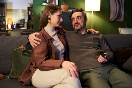 Couple sitting closely on sofa, embracing and talking while holding remote control, showing relationship bonding. Ideal for family lifestyle, home leisure, streaming ads