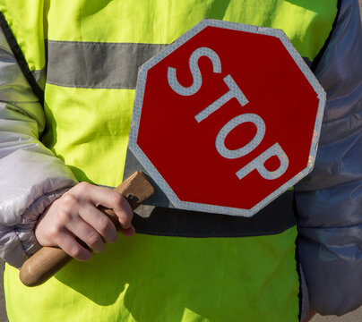 A crossing guard wearing a high visibility reflective yellow jacket firmly grips the wooden handle of a hexagonal red and white warning sign to signal for traffic to halt
