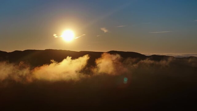 Streak of golden mist drifts below a dark mountain ridge at sunrise, sun glowing in upper frame against a gradient blue sky, lens flare crossing the scene