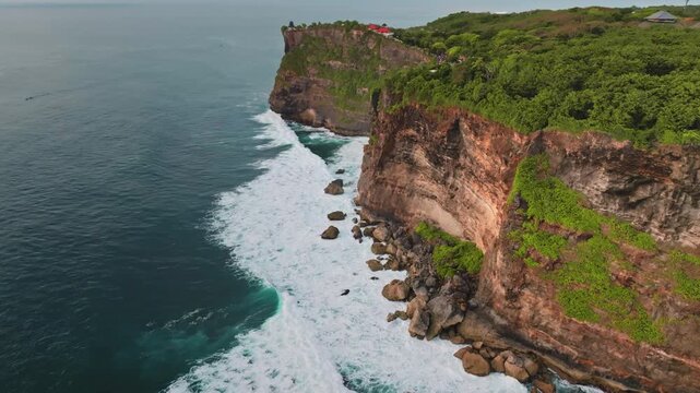 Towering sea cliff facing the vast Indian Ocean, showcasing powerful waves breaking against the rocky coastline of the tropical Uluwatu region on the stunning Bukit Peninsula. Aerial view drone flight